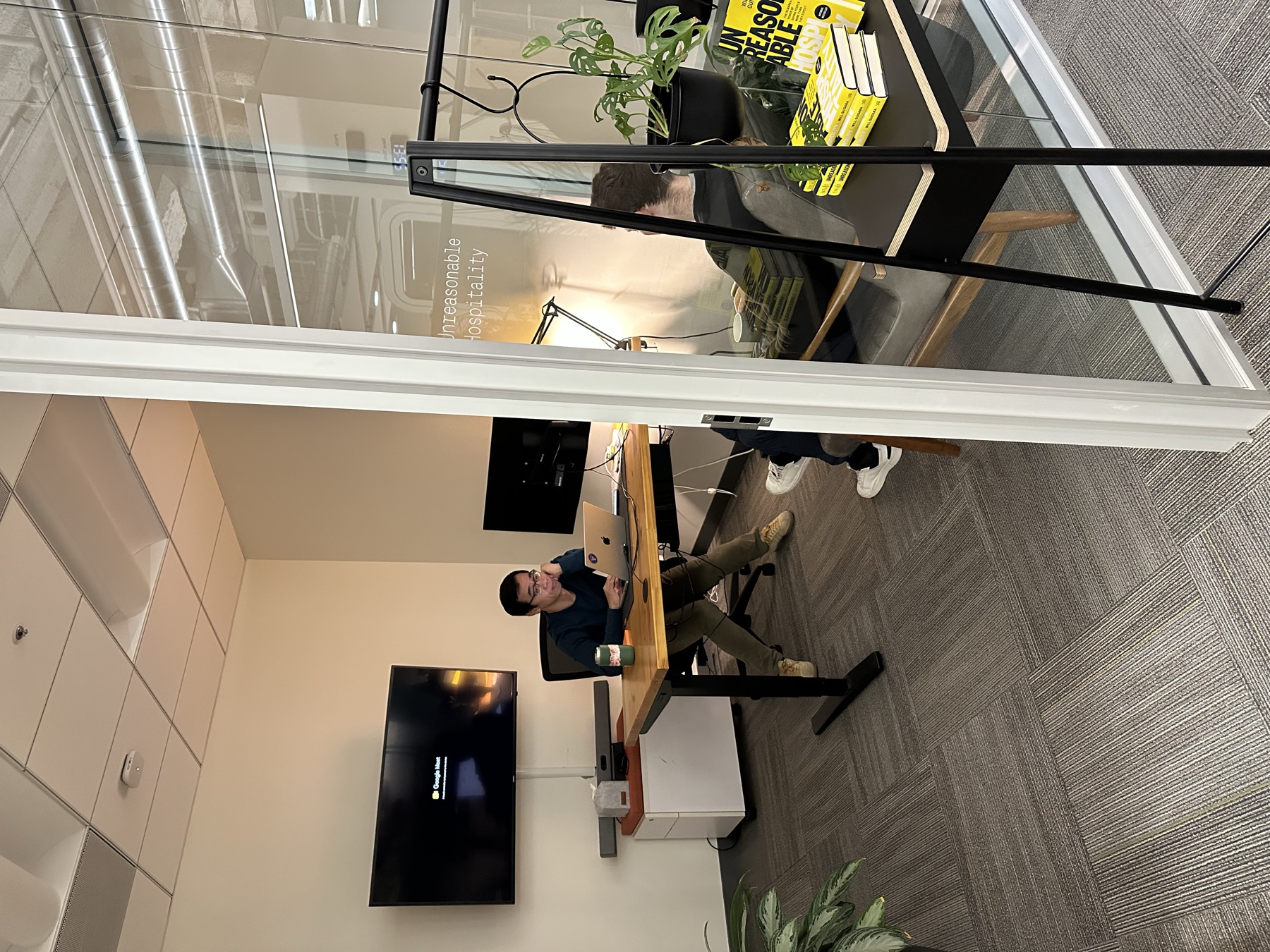 A person working at a laptop inside a glass-walled voice room while a colleague sits nearby.